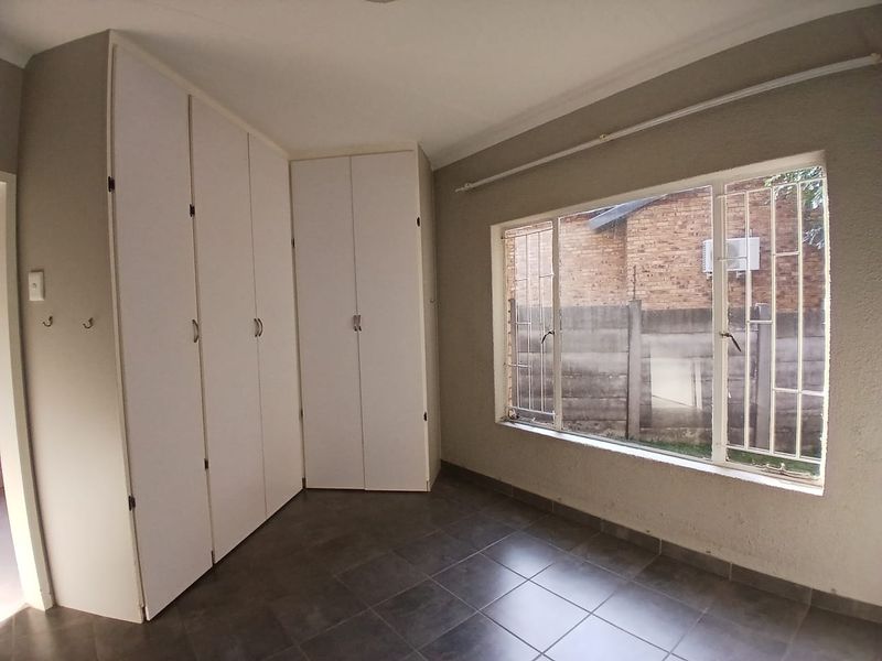 A bright, empty bedroom featuring tiled flooring, built-in cupboards, and large windows fitted with security bars.