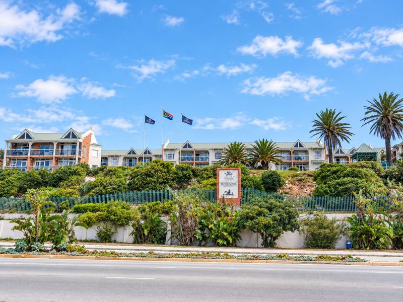 Street-level view of the Brookes Hill apartment complex, featuring multi-story residential buildings with balconies, landscaped gardens, and secure perimeter fencing.