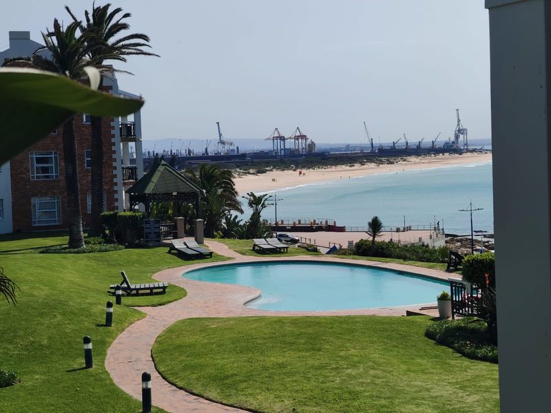 A well-maintained communal swimming pool area set within landscaped gardens, featuring a paved walkway and gazebo, overlooking the beach and harbor.