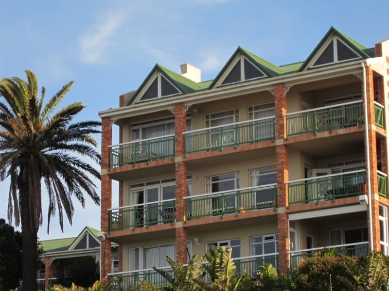 A multi-story apartment building featuring a brick facade, green metal roofing, and private balconies with metal railings for each unit.