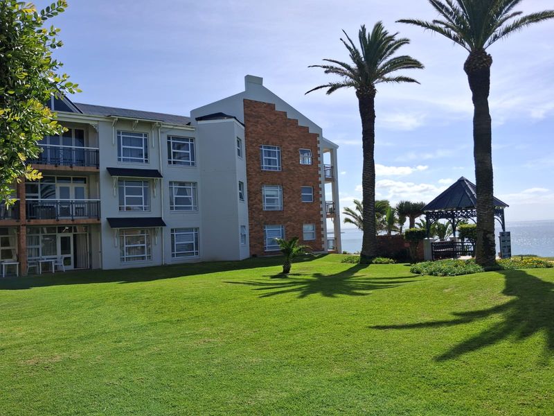 A view of the apartment building's rear facade featuring brick detailing, private balconies, and a well-maintained lawn with a gazebo overlooking the ocean.
