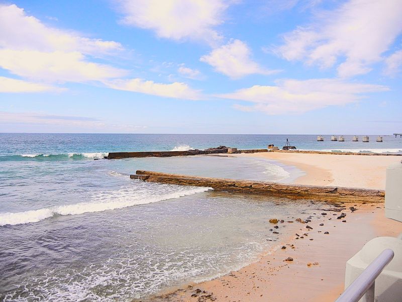 A scenic coastal view featuring a sandy beach, ocean waves, and stone breakwaters under a bright blue sky.