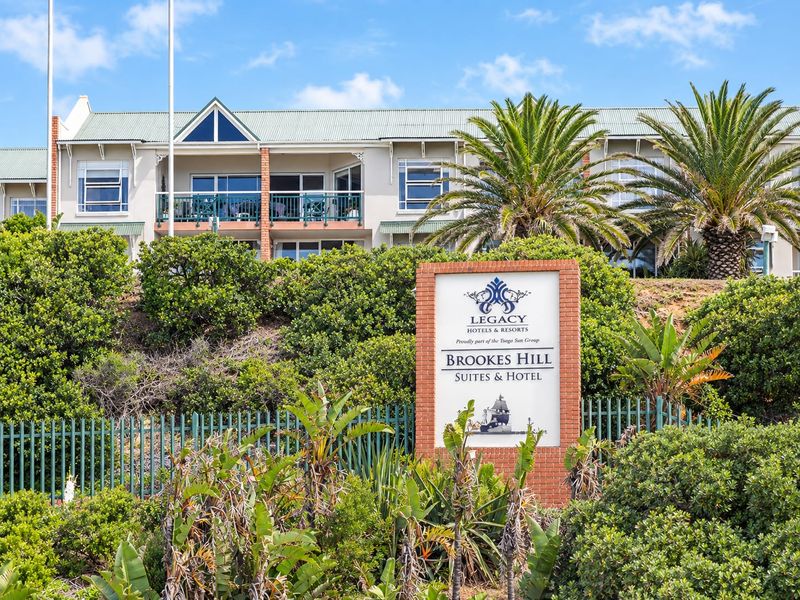 Exterior view of the Brookes Hill Suites and Hotel complex, featuring landscaped gardens, security fencing, and multi-level apartment balconies.