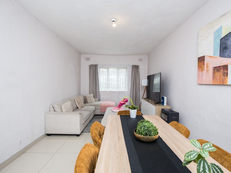 Open-plan living and dining area featuring tiled flooring, neutral walls, and a window with security bars, offering a functional space for daily living.