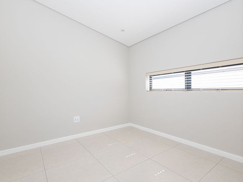 A clean, neutral-toned bedroom featuring large format floor tiles, a horizontal window with fitted blinds, and recessed lighting.