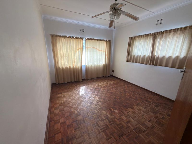 An empty bedroom featuring original parquet flooring, a ceiling fan, and large windows with neutral curtains.