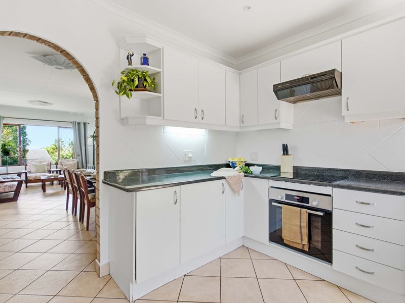 A clean, functional kitchen featuring white cabinetry, dark stone countertops, built-in oven, and hob, connected to the living area via a charming brick archway.