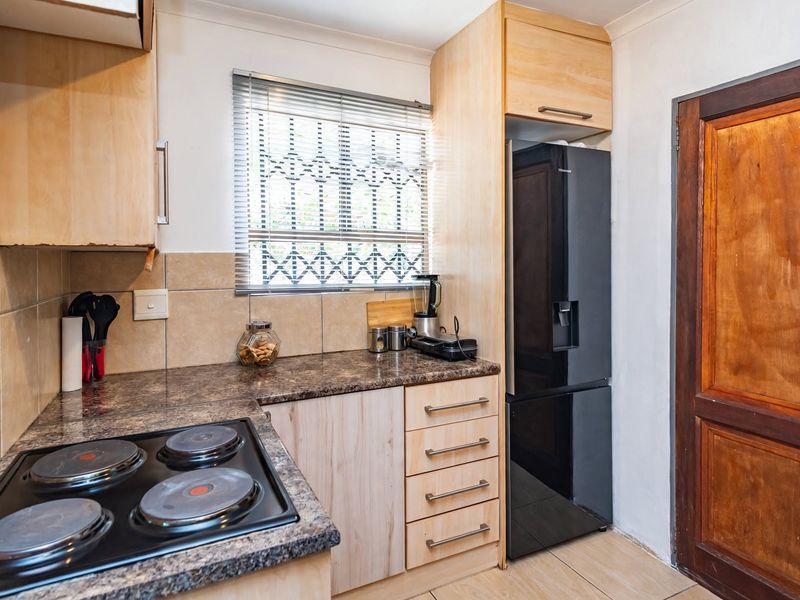 Compact kitchen featuring light wood-finish cabinetry, granite-look laminate countertops, an electric hob, and tiled backsplash with a window security gate.