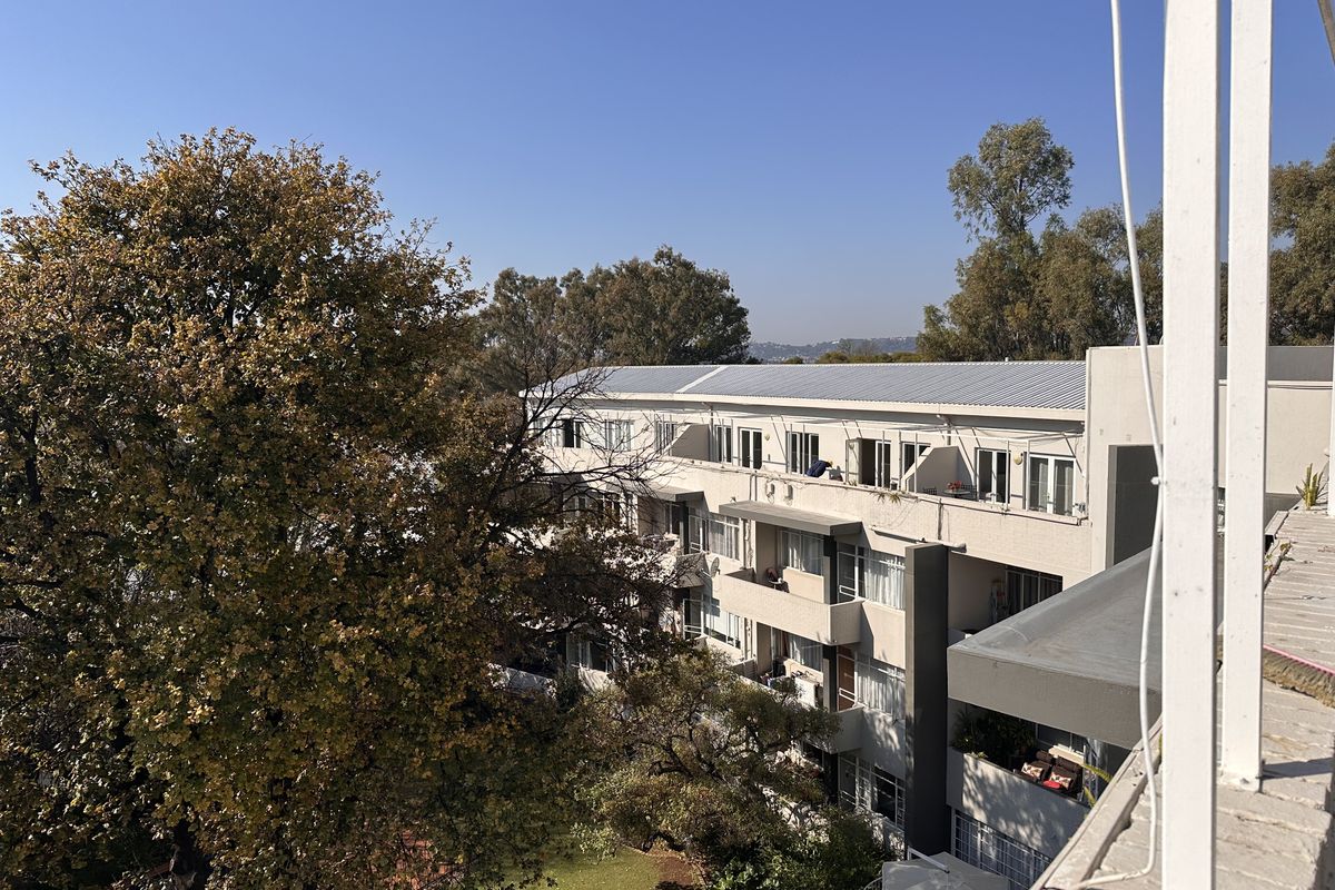 An elevated view of the apartment complex rear facade, featuring multiple balconies, sliding doors, and a metal roof set against a landscaped garden.