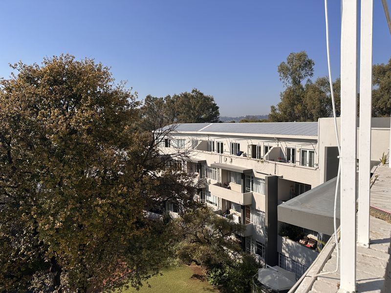 An elevated view of the apartment complex rear facade, featuring multiple balconies, sliding doors, and a metal roof set against a landscaped garden.