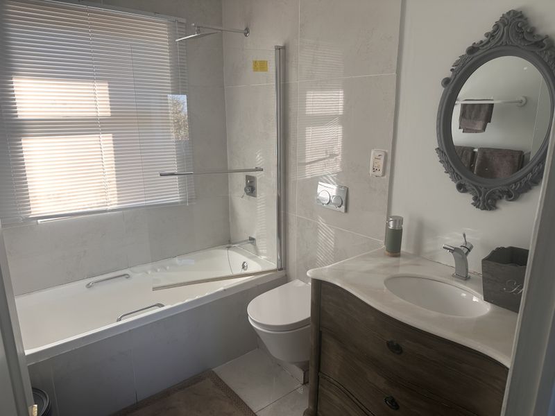Modern bathroom featuring a wall-mounted toilet, built-in bathtub with glass screen, rain shower head, and a vanity cabinet with integrated sink.