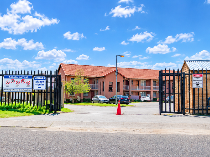View of the apartment complex entrance featuring secure access control gates, a guard house, and paved driveway access to the residential buildings.