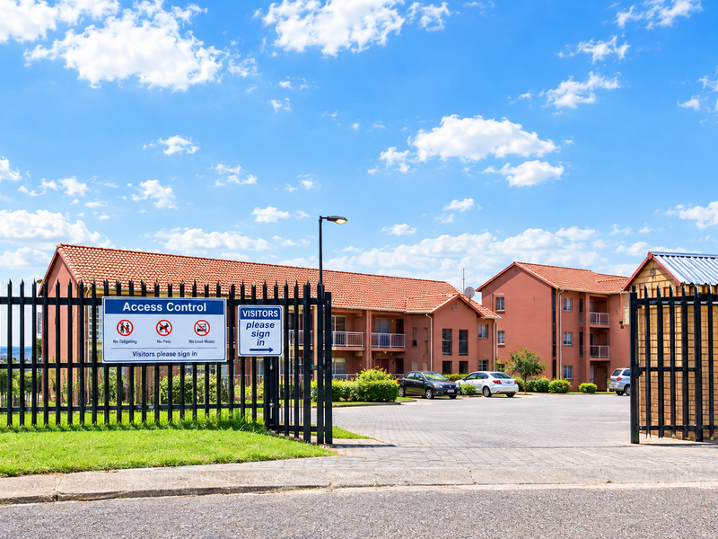 Front entrance view of an apartment complex featuring secure gated access, paved driveway, and multi-story residential buildings with terracotta tiled roofs.