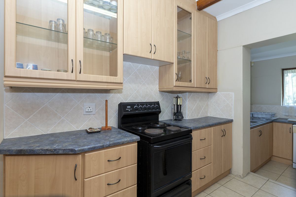 A functional kitchen featuring light wood-finish cabinetry, dark laminate countertops, a freestanding electric stove, and tiled backsplash, with an adjacent scullery area.
