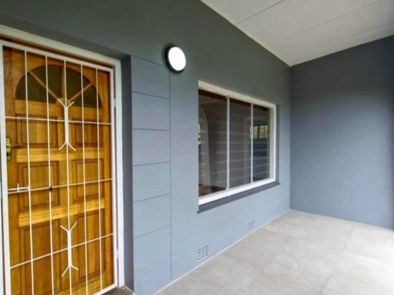 A clean, covered patio area featuring tiled flooring, a secure wooden entrance door with a security gate, and a large sliding window.