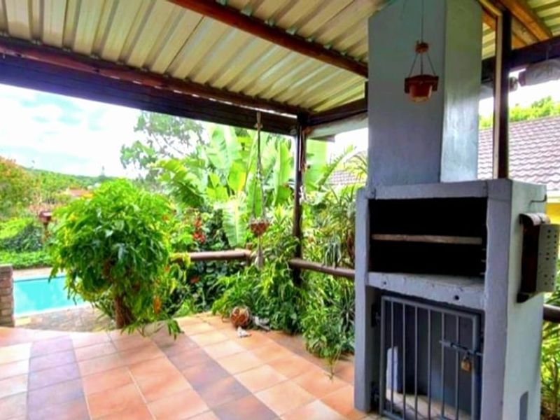 Covered patio area featuring a built-in braai, tiled flooring, and timber railings, overlooking the garden and swimming pool.