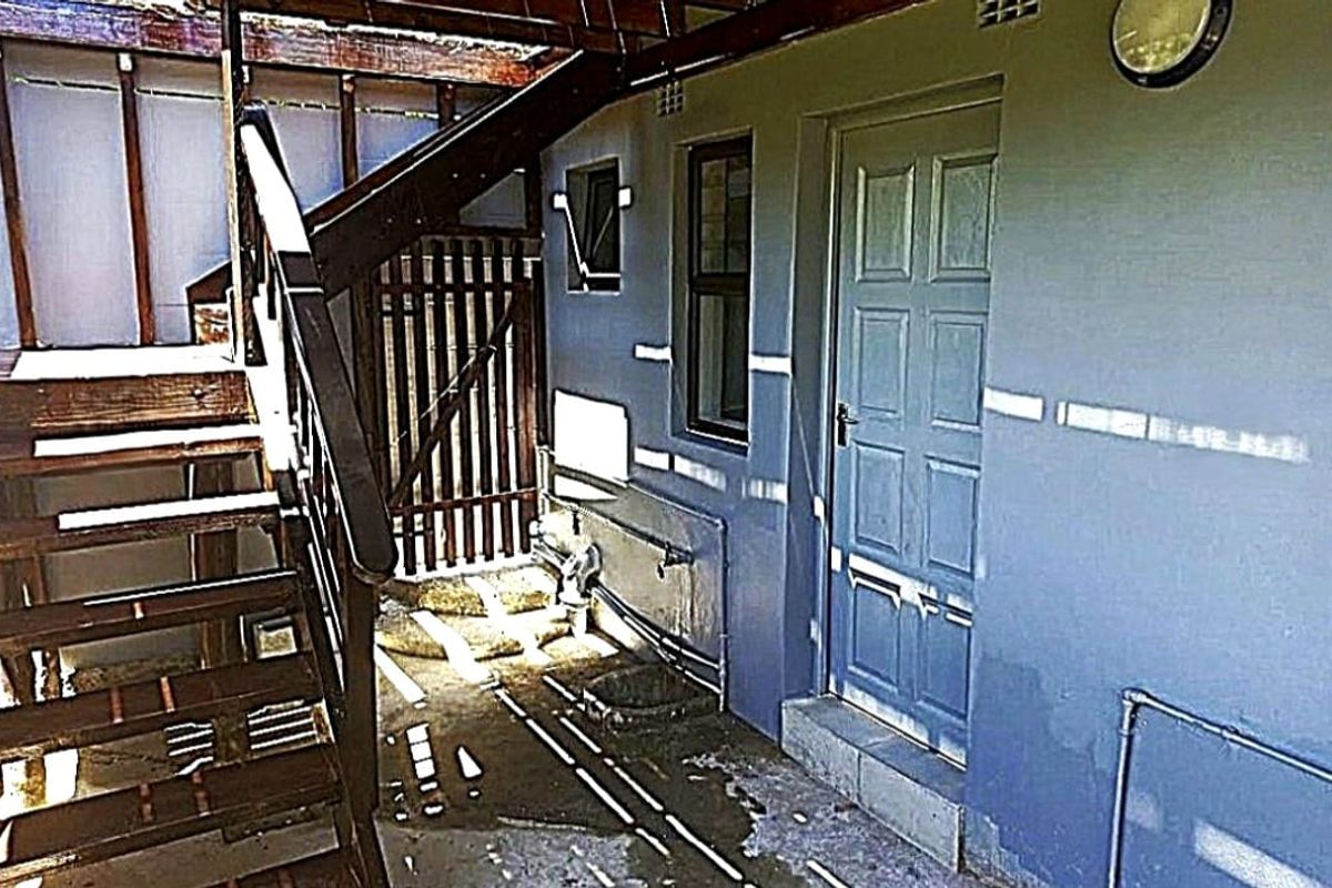 Rear exterior area featuring a timber staircase, external door, and utility access, showing signs of significant weathering and wear.