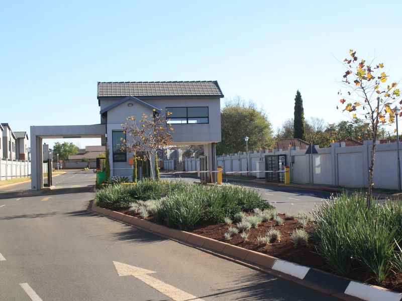 A well-maintained residential estate entrance featuring a security gatehouse, boom gate access, perimeter walling, and landscaped garden beds along the paved driveway.