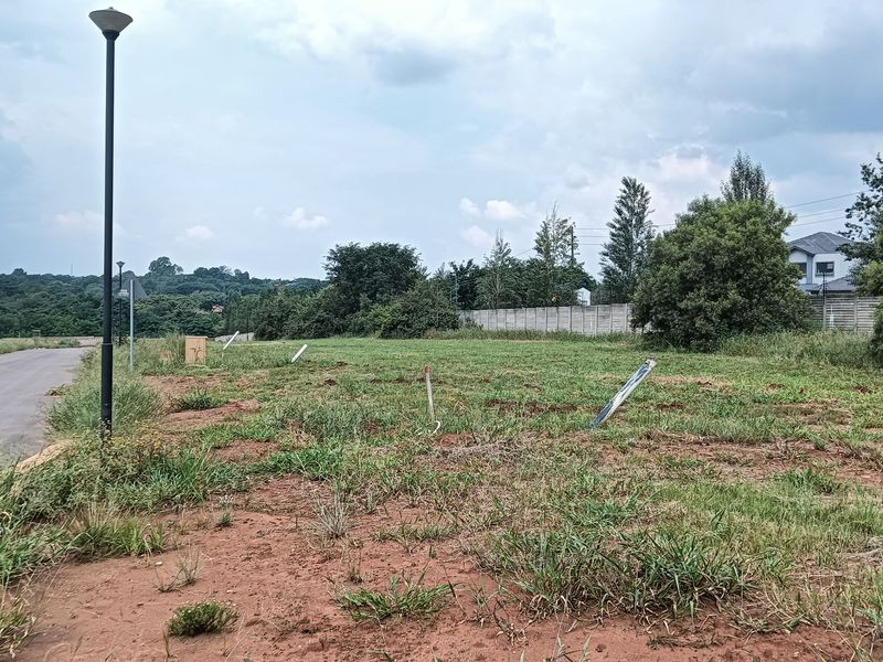 A vacant residential plot of land featuring grass cover, adjacent to a paved road with street lighting and perimeter boundary walling.