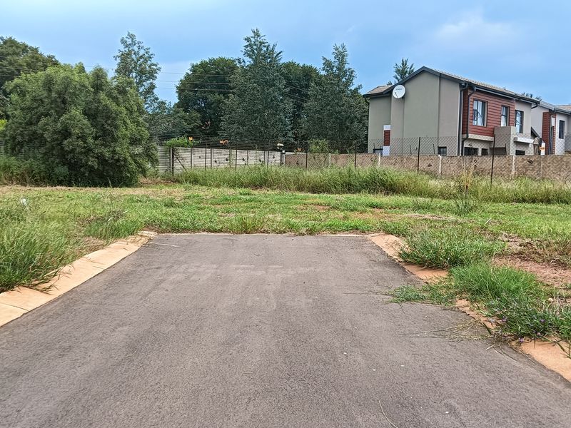 A vacant residential plot featuring a paved driveway entrance and perimeter fencing, situated in a developed neighborhood setting.