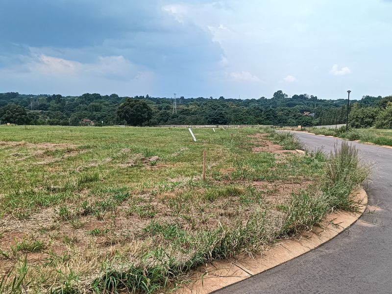 A vacant residential plot featuring a paved road frontage and established survey markers, ready for development in a scenic, green setting.