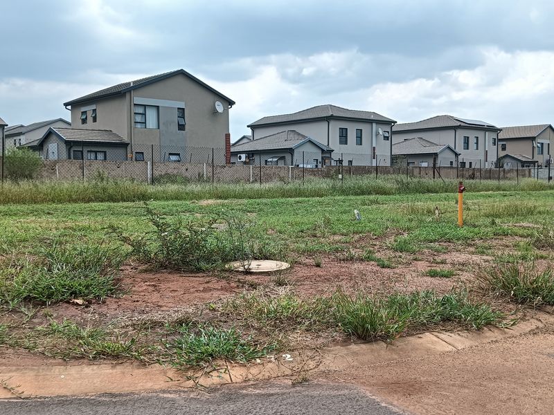 A vacant residential plot of land featuring grass cover, a manhole, and a fire hydrant, situated in a developed suburban neighborhood.