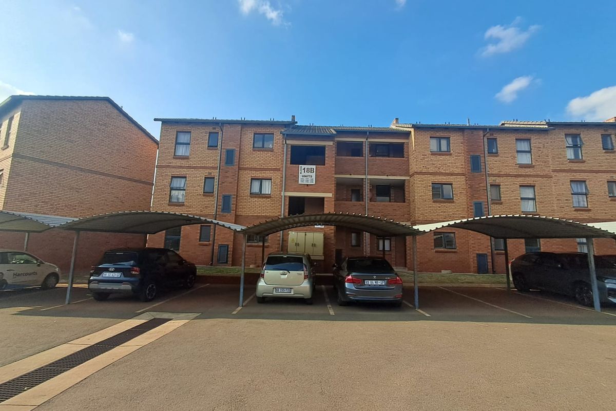 Front elevation of a multi-story brick apartment complex featuring covered parking bays and a paved driveway area.