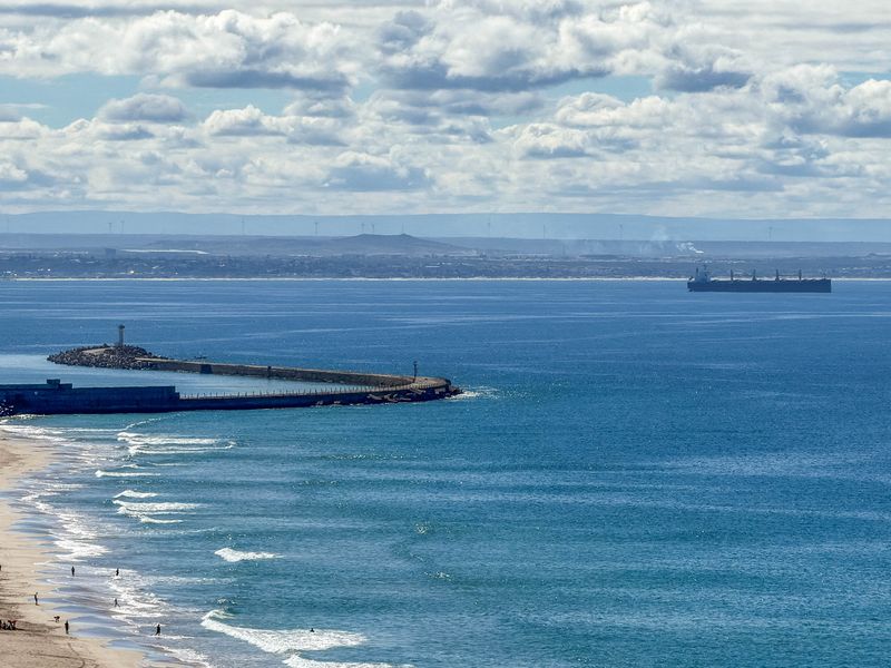 A scenic elevated view of the coastline, featuring a sandy beach, a breakwater pier, and the open ocean with distant shipping vessels.