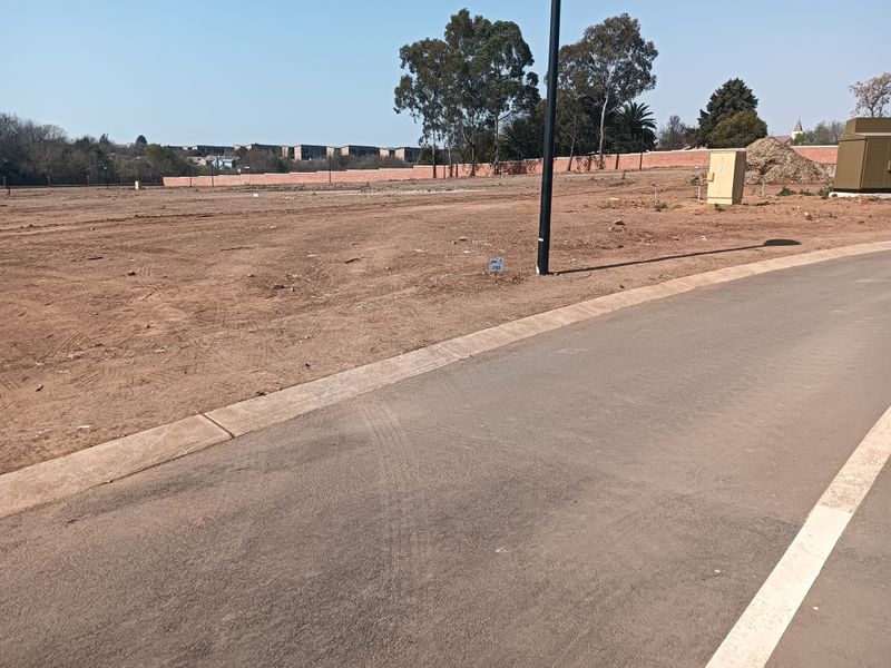 A vacant residential plot featuring a paved road frontage, concrete curbing, and established boundary walls in a developed estate setting.