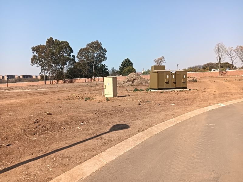A vacant residential plot featuring a paved road frontage, perimeter wall, and installed utility infrastructure including electrical distribution boxes.