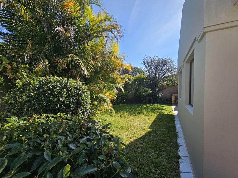 A private rear garden featuring a well-maintained lawn, mature landscaping, and a built-in brick braai area adjacent to the townhouse exterior.