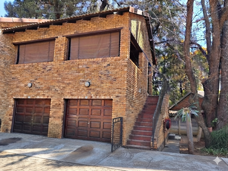 A two-story face brick apartment building featuring a double garage, external staircase with metal railing, and mature surrounding trees.