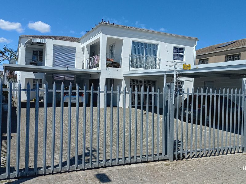 Front elevation of a two-story flat property featuring a paved driveway, carport, and secure metal perimeter fencing.