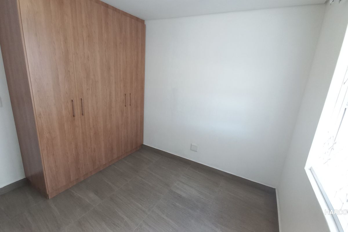A clean, empty bedroom featuring modern wood-finish built-in wardrobes, neutral tiled flooring, and a window fitted with a security gate.