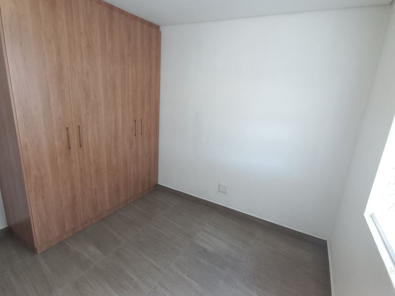 A clean, empty bedroom featuring modern wood-finish built-in wardrobes, neutral tiled flooring, and a window fitted with a security gate.