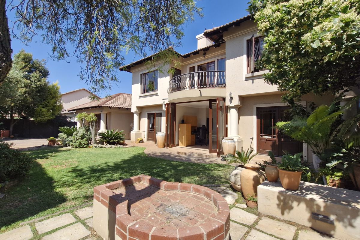 A well-maintained rear exterior view featuring a landscaped garden, paved patio area with a brick fire pit, and a balcony with wrought iron railings.