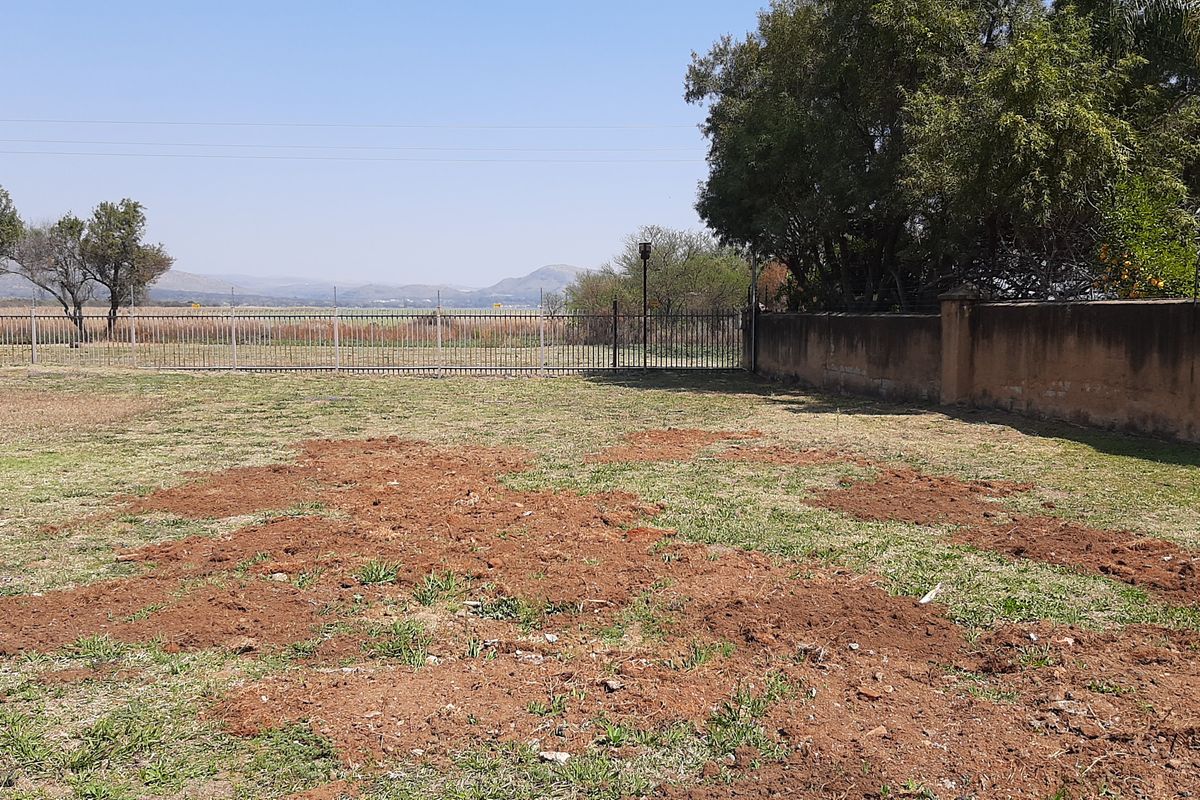 A vacant residential plot featuring perimeter metal fencing, a boundary wall, and mature trees with views of the surrounding landscape.