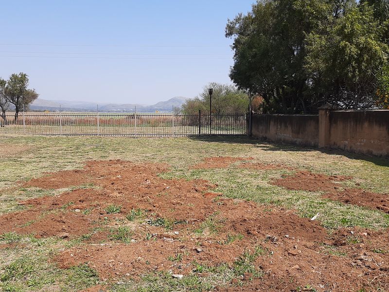 A vacant residential plot featuring perimeter metal fencing, a boundary wall, and mature trees with views of the surrounding landscape.