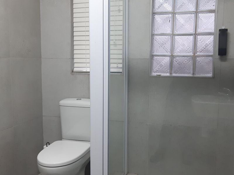 A clean, functional bathroom featuring a modern toilet, a glass-enclosed shower, and neutral wall tiling with a glass block window for natural light.