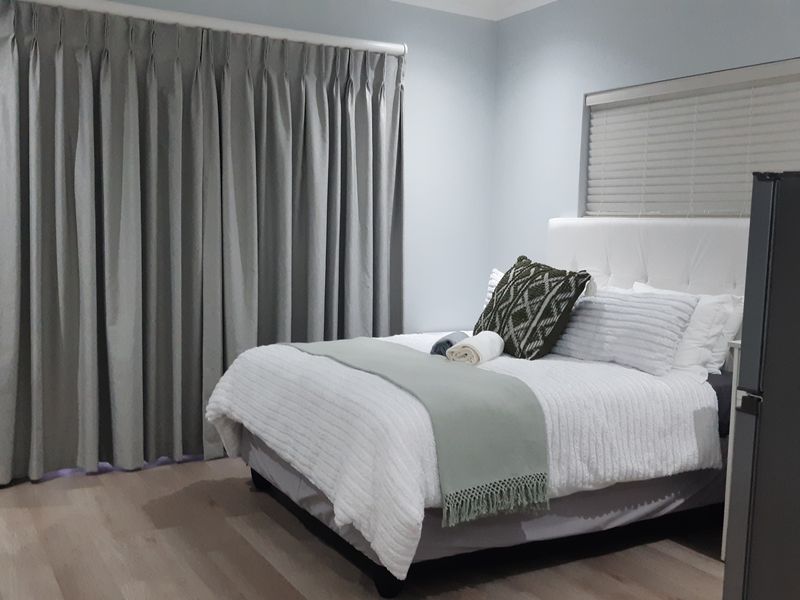 A well-maintained bedroom featuring light-toned laminate flooring, neutral wall finishes, and fitted window treatments including curtains and venetian blinds.