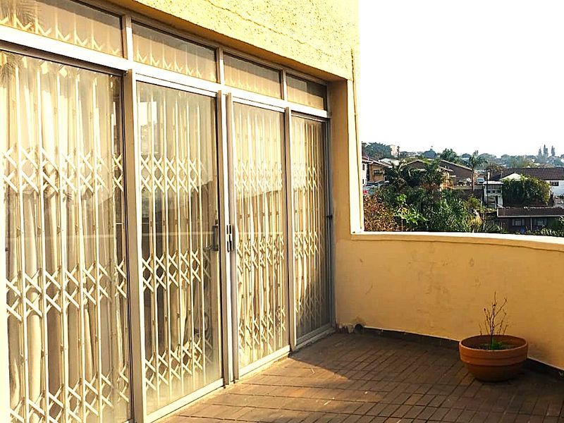 A spacious balcony featuring tiled flooring, sliding glass doors with security gates, and a parapet wall with views of the surrounding neighborhood.