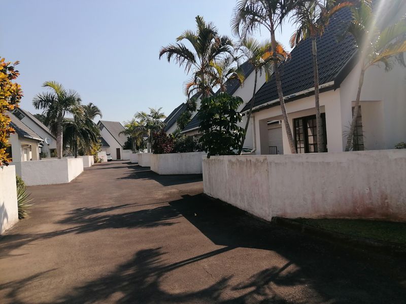 A view of the townhouse complex driveway featuring paved access, boundary walls, and established palm trees leading to the residential units.
