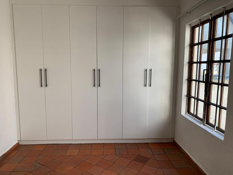 A bright bedroom featuring extensive built-in white cabinetry, traditional terracotta tile flooring, and a window fitted with security bars.