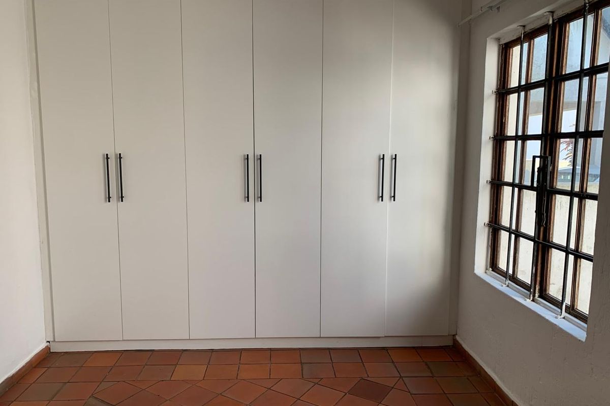 A bright bedroom featuring extensive built-in white cabinetry, traditional terracotta tile flooring, and a window fitted with security bars.