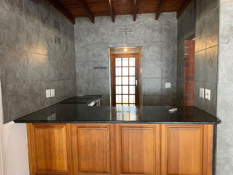A kitchen area featuring wooden cabinetry, granite countertops, exposed timber ceiling beams, and a tiled splashback with a rear access door.