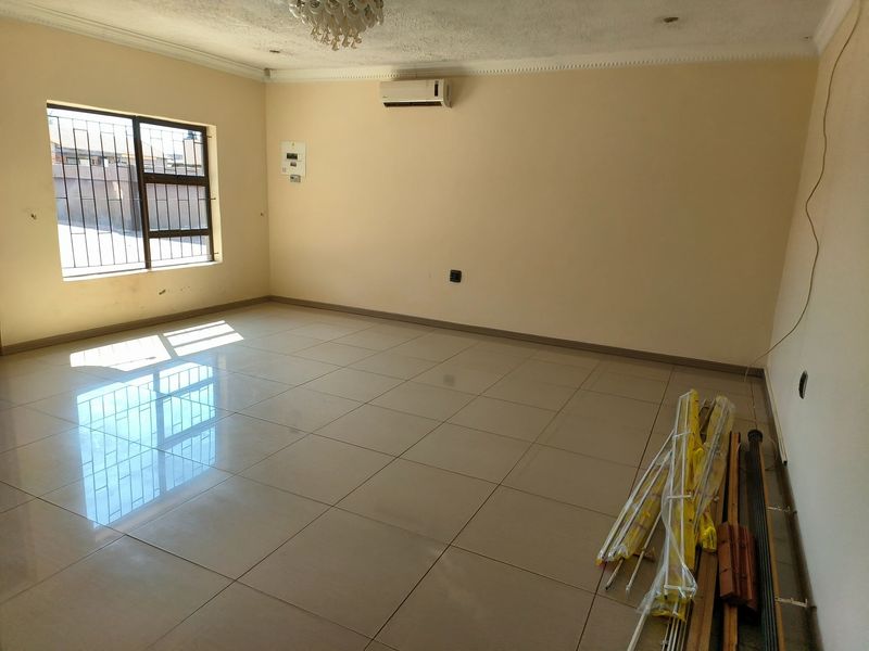 A spacious, empty living room featuring neutral tiled flooring, a wall-mounted air conditioning unit, and security bars on the window.