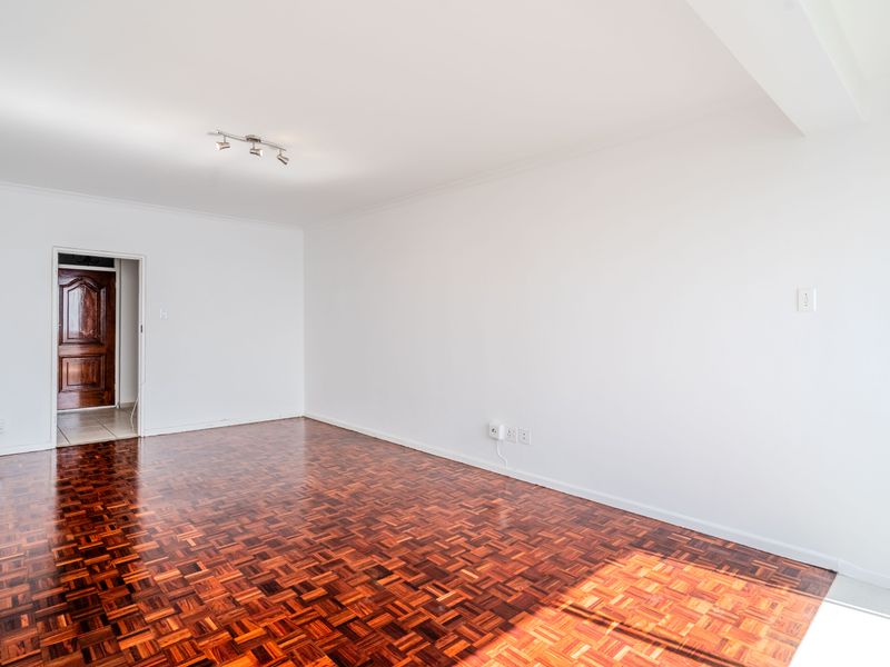 A bright, empty living room featuring polished parquet flooring, white walls, and a ceiling-mounted spotlight fixture.