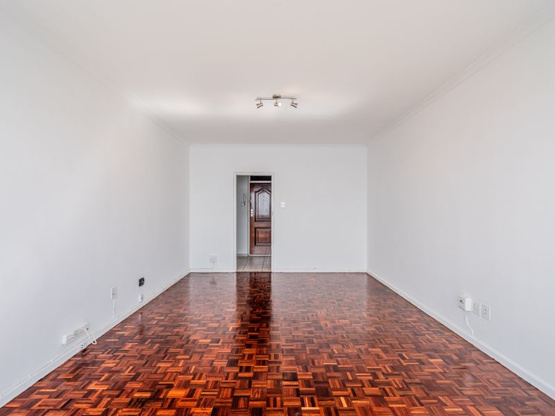 A bright, empty living room featuring polished parquet flooring, neutral white walls, and a ceiling-mounted spotlight fixture.