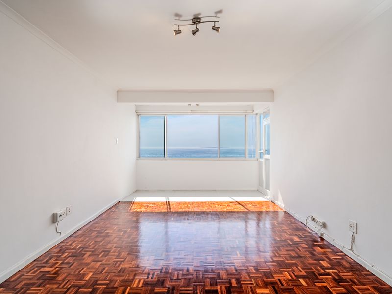A bright, empty living room featuring polished parquet flooring and a large window offering expansive ocean views.