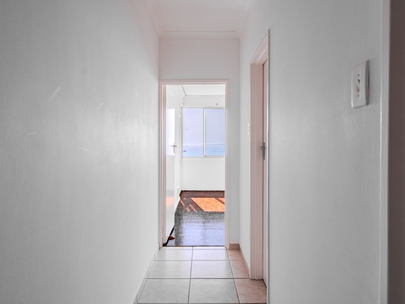 A simple, neutral hallway with tiled flooring leading towards a sunlit room with a view.